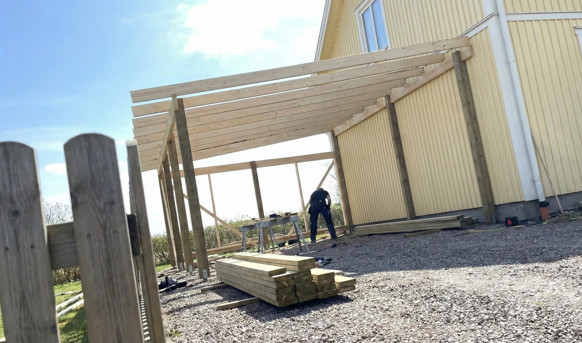 Puinen autokatoksen runko rakenteilla keltaisen talon vieressä aurinkoisena päivänä. Maassa on pinottuna rakennuspuuta ja työntekijä näkyy taustalla.