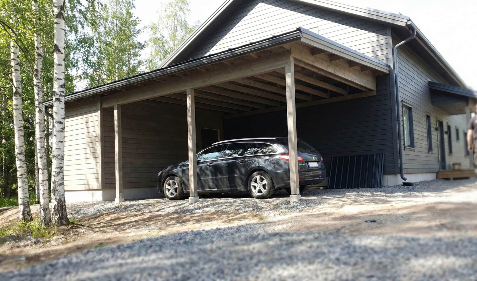 Musta henkilöauto on pysäköity uuden näköisen talon tummanharmaaseen autokatokseen. Koivuja reunustaa pihaa aurinkoisena päivänä.